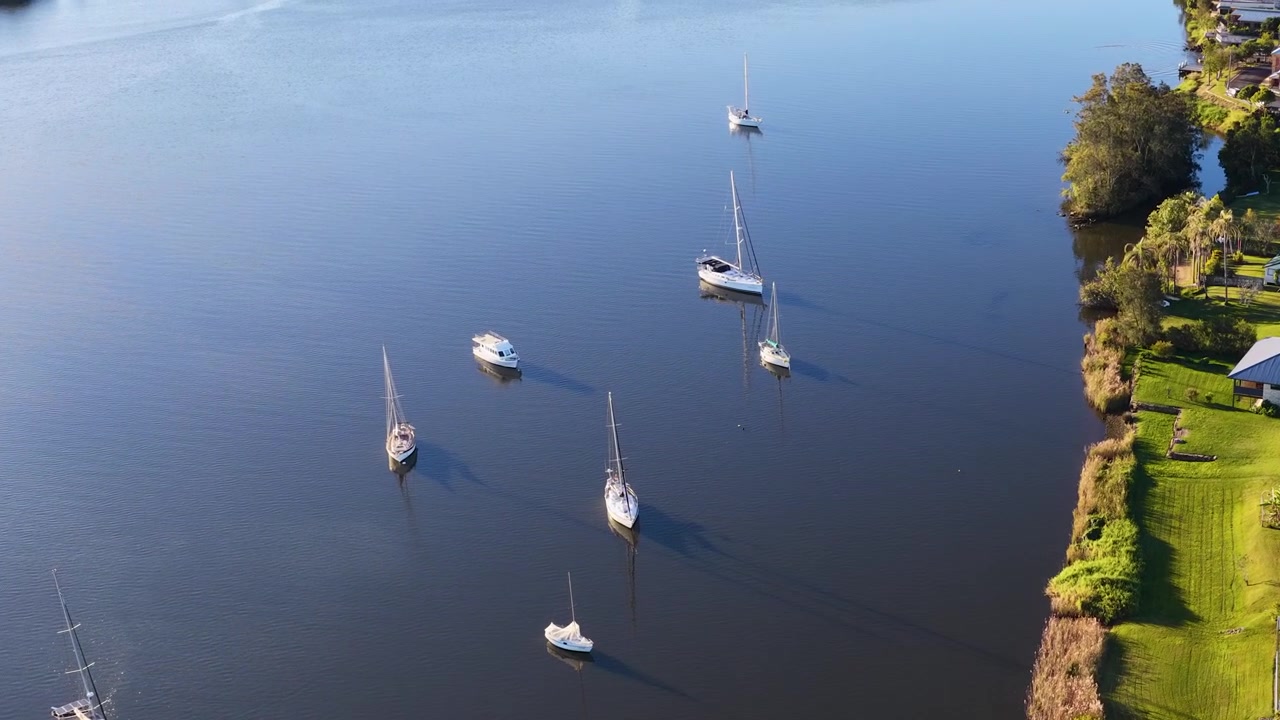 Drohnenaufnahme aus der Vogelperspektive von Segelbooten, die auf ruhigem Flusswasser in der Nähe von Vorstadthäusern in Maclean vertäut sind.