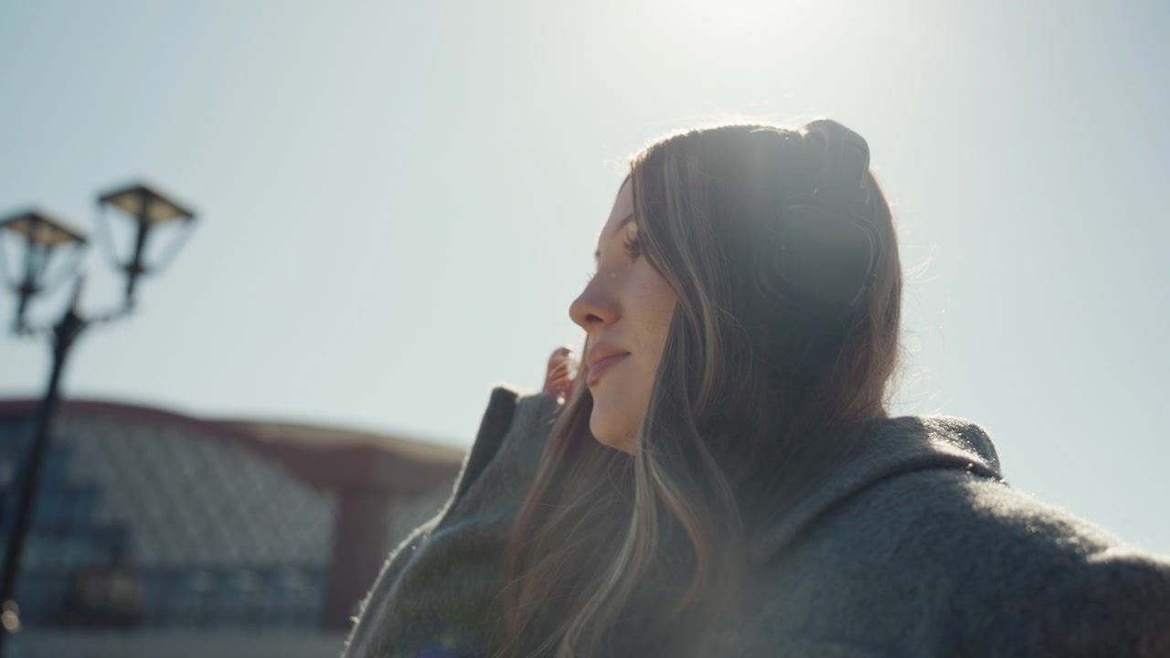 Woman with headphones. Cheerful woman in cozy coat smiling. Candid shot of woman adjusting headphones outdoors. Playful woman with rosy cheeks and headphones under sunlit station backdrop