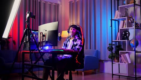 asian boy streamer smiling to camera while playing game over network on personal computer. live stream video game, desk illuminated by rgb led strip light