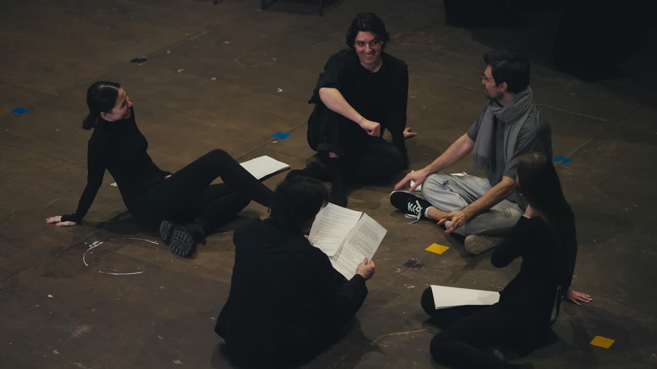 검은색 의상을 입은 자신감 넘치는 젊은 배우들이 감독과 함께 무대에 앉아 극장 무대에서 연극의 시나리오를 가진 종이를 손에 들고 있습니다.