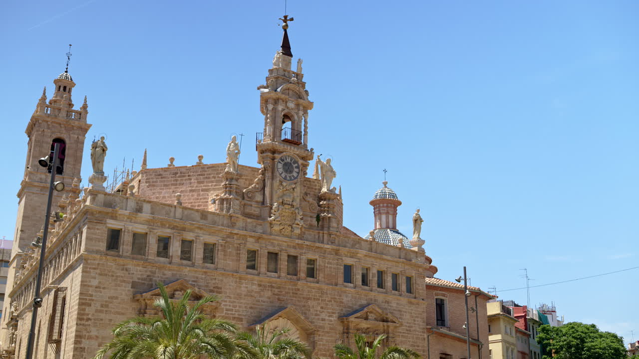 Low angle view of the baroque church Sant Joan del Mercat with statues, central clock and belfry, and blue-tiled domes against a bright blue sky in Valencia, Spain
