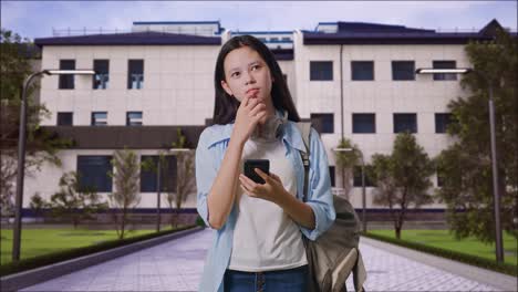estudiante adolescente asiática con una mochila usando un teléfono inteligente y pensando mientras está de pie frente a un edificio escolar