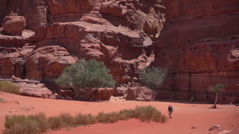 Hombre-Solitario-Caminando-Sobre-La-Arena-Del-Desierto-Bajo-Escarpados-Acantilados-De-Arenisca-Roja-En-Un-Día-Caluroso-Y-Soleado