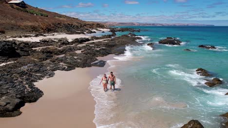 A-couple-walk-hand-in-hand-along-a-white-sand-beach-as-the-blue-ocean-water-rolls-over-their-feet-on-the-shore