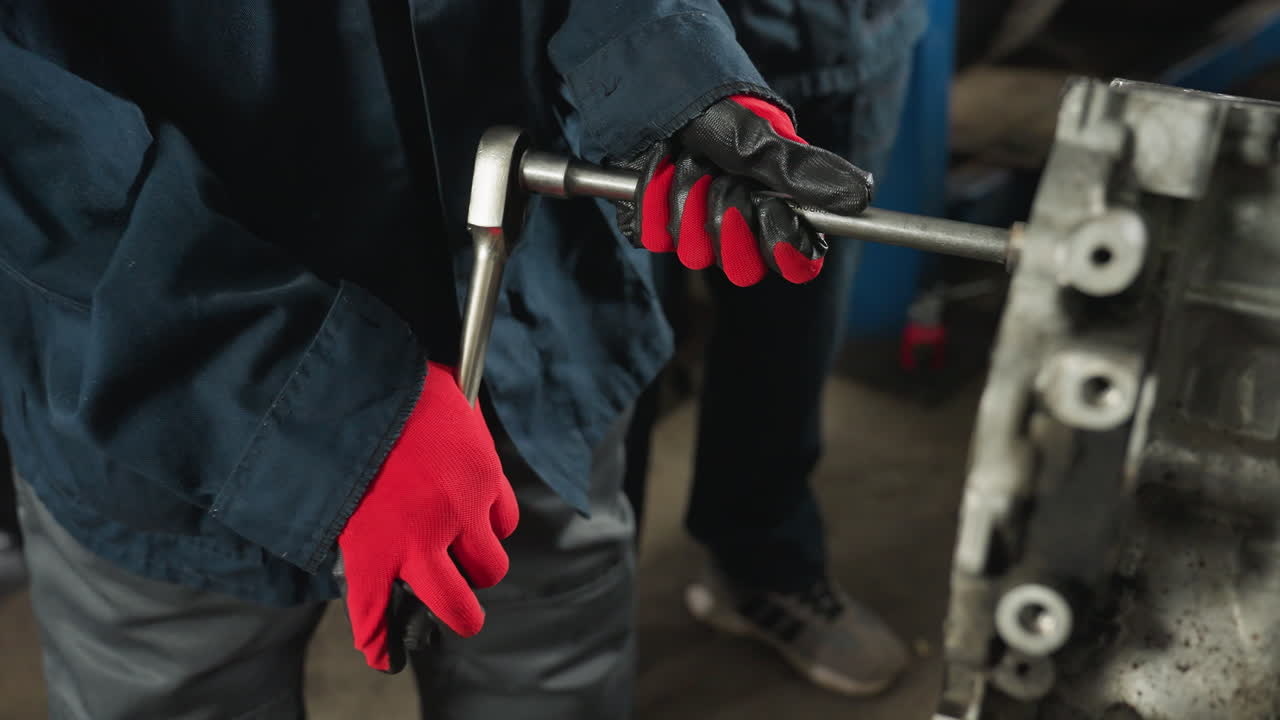 primer plano de un mecánico con guantes rojos apretando pernos en el bloque del motor, con las piernas de otra persona visibles, trabajando juntos en un taller ocupado