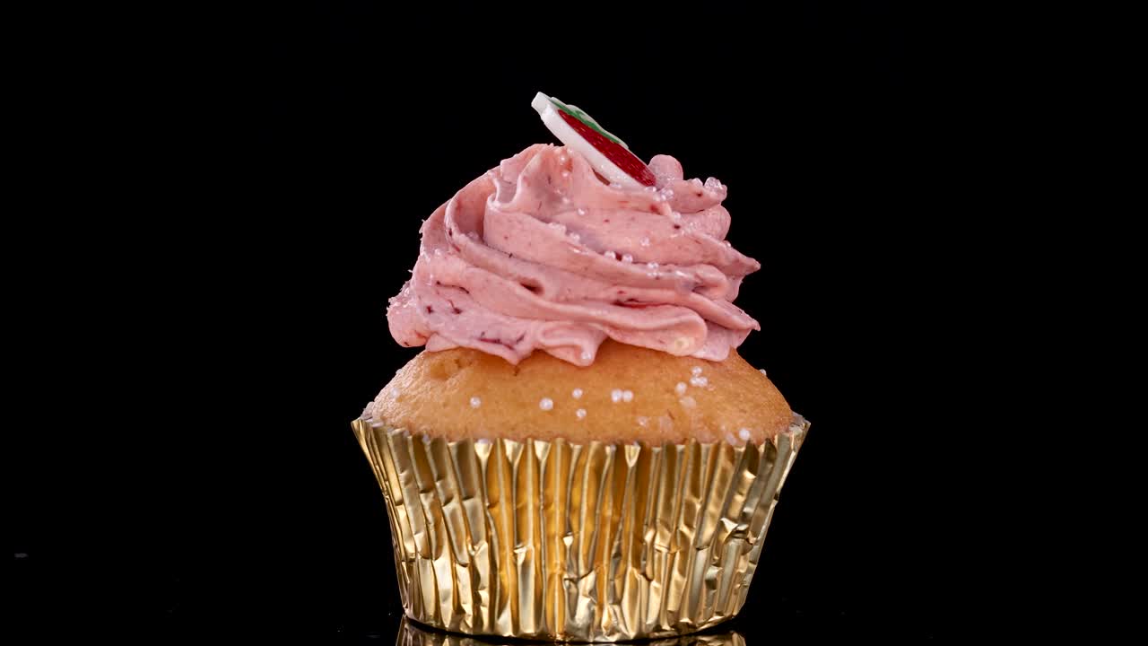 A single gourmet cupcake with pink strawberry frosting and gold wrapper rotates smoothly against a seamless black background, illuminated by even studio lighting