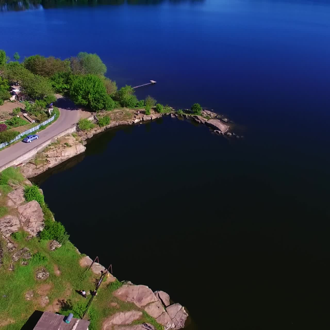 Automobile road with some cars at the waterfront. Drone flying over the azure river on sunny day. Agricultural fields at backdrop