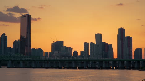 train crossing han river moving on railway bridge, seoul yeouido island skyline with 63 building in silhouette during picturesque orange sunset, south korea