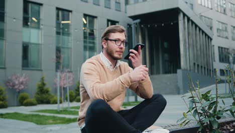 successful caucasian man in glasses using cellphone while sitting crossing legs on bench of city financial district. outdoors, man typing online sms on his phone sitting on a bench