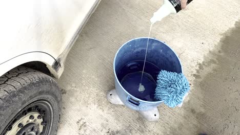 pouring a little bit of car soap shampoo into a bright blue bucket with a microfiber washing mitt on top and a filthy white car with all terrain tires next to it