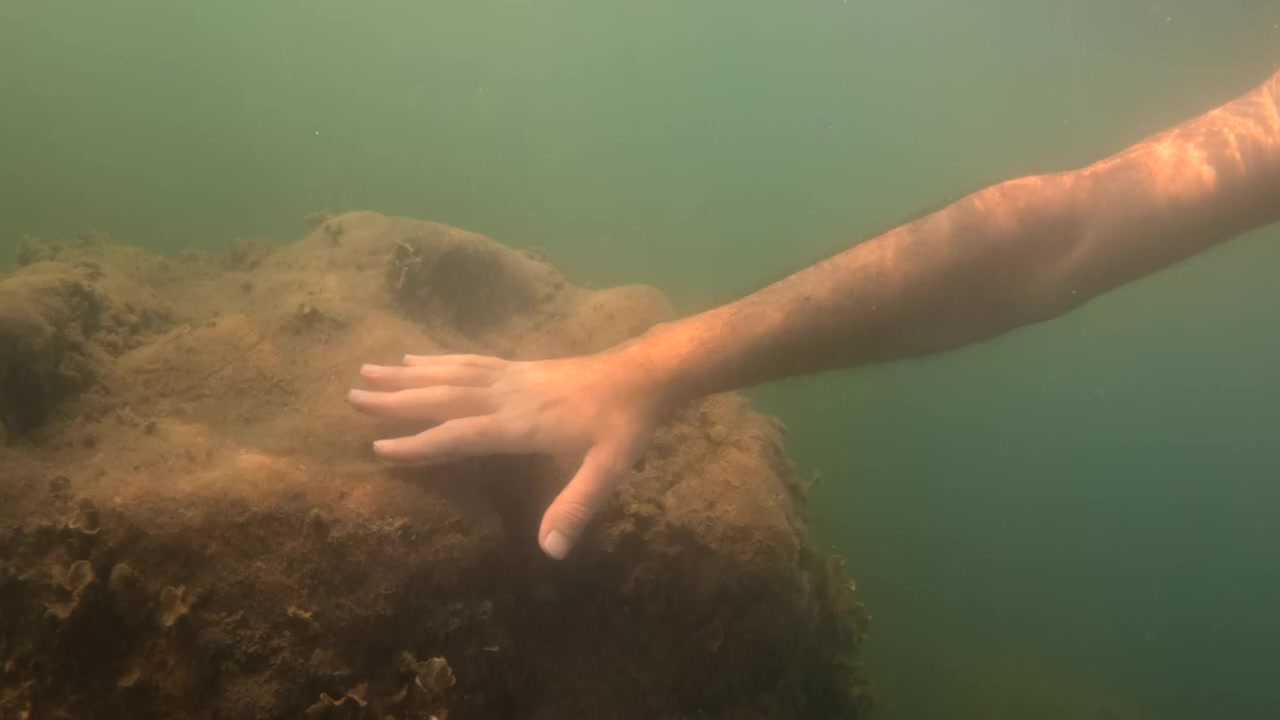 Mand svømmer under vandet, iført gule svømmebriller, med sollys der reflekteres på vandoverfladen og skaber mønstre