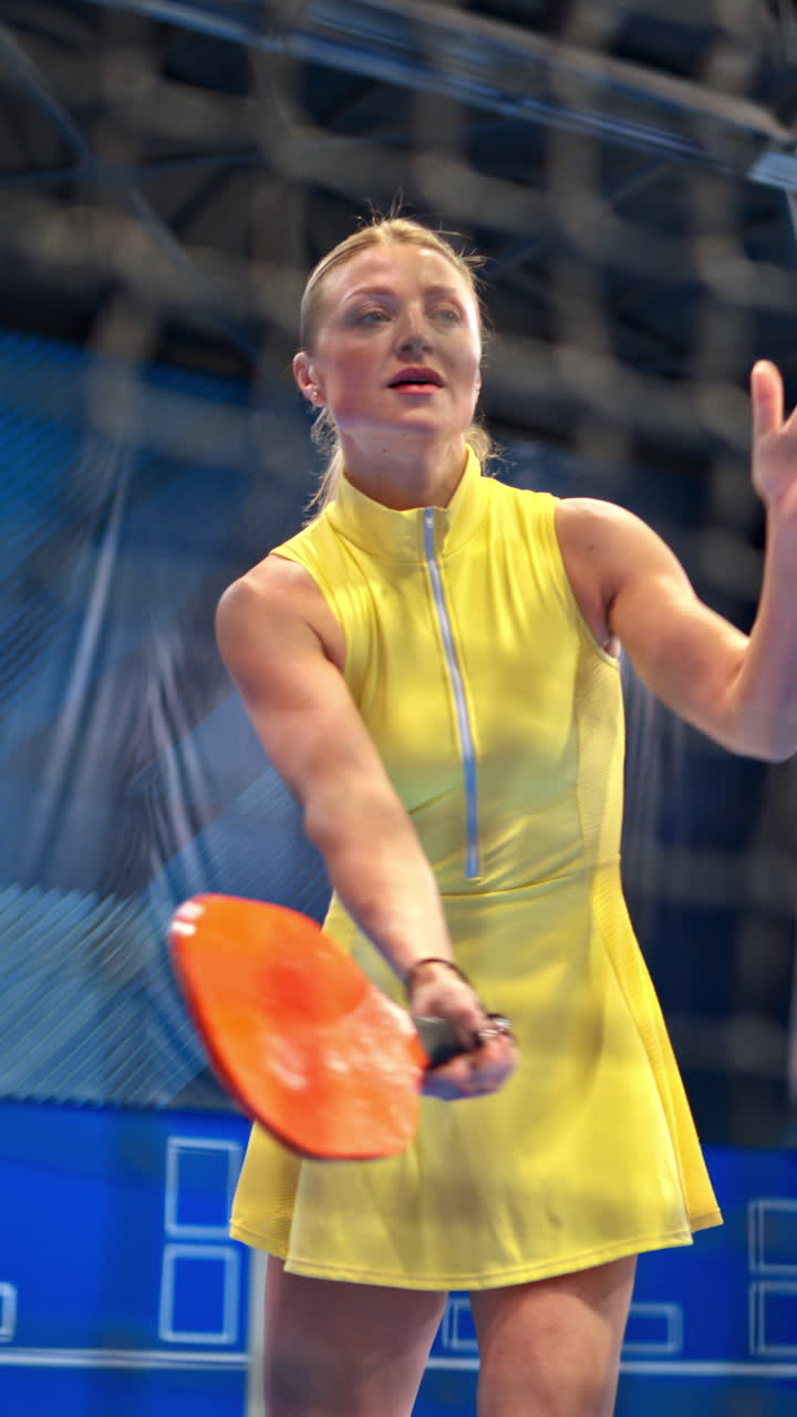 Woman in a yellow dress training to play pickleball on a blue, inside court. Vertical