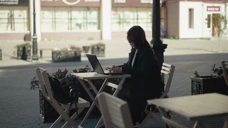 student working on laptop outdoors at mall surrounded by wooden chairs, plants, and tripod, seated at table with coffee cup, paved walkway, bird flying down in the distance