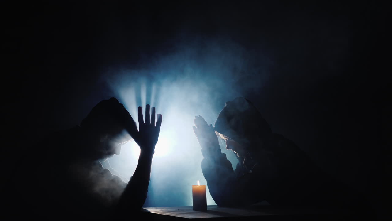 dos personas se sientan en una mesa rezando para que una vela se queme cerca de ellos detrás de la niebla refrescada por un foco