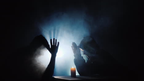 two people sit at a table praying a candle burns near them behind the mist refreshed by a spotlight