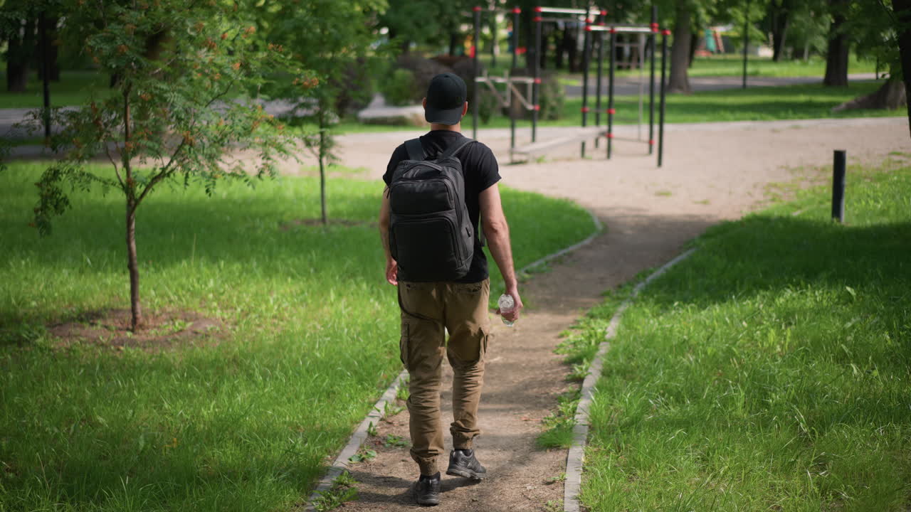 Individual Walks Along City Park Trail, Traveler Navigates City Greenery With Determined Steps, Person Advances Purposefully Through Urban Park Environment Carrying Backpack And Energy