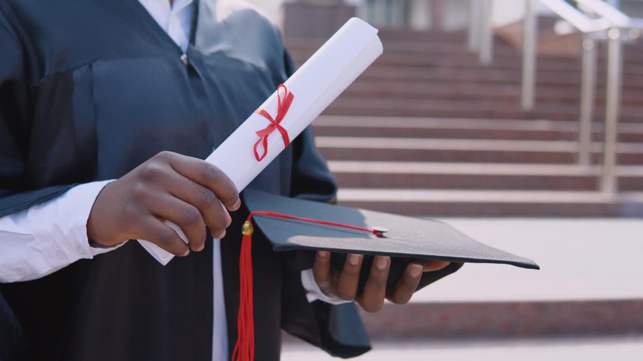 sombrero de maestro cuadrado y diploma de graduación en las manos de una mujer afroamericana graduada de la universidad en el fondo de las escaleras desde afuera.