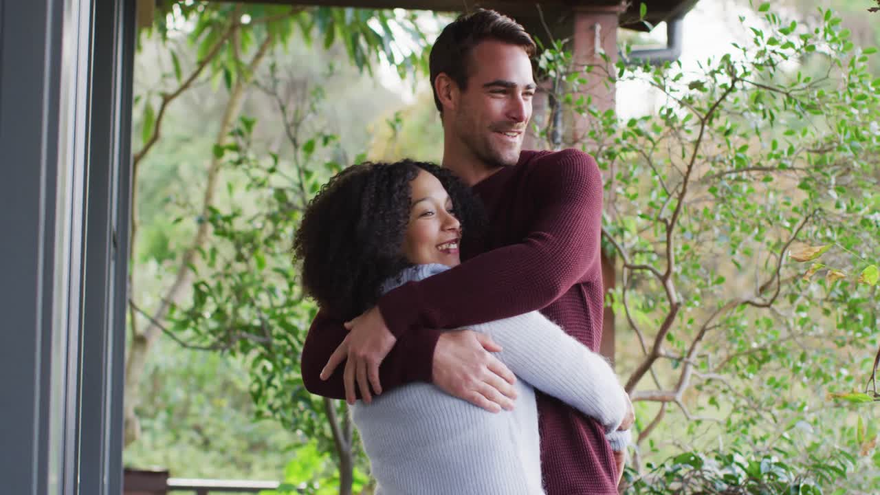 pareja feliz diversa abrazando y riendo en la sala de estar, mirando por la ventana en el campo