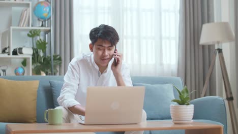 Smiling Asian Man Answering The Call On Smartphone While Using Laptop Computer In The Living Room