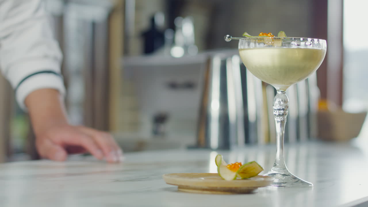 Close-up of a decorated cocktail glass next to an appetizer, with a bartender adjusting the items on the bar