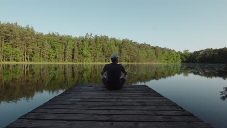 Man-sits-cross-legged-at-end-of-wooden-dock-pier-looking-out-in-wonder-at-Lac-de-Lispach