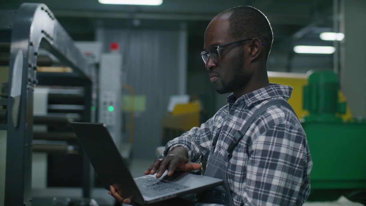 Black Engineer Using Laptop to Program Automated Machinery in Printing Factory