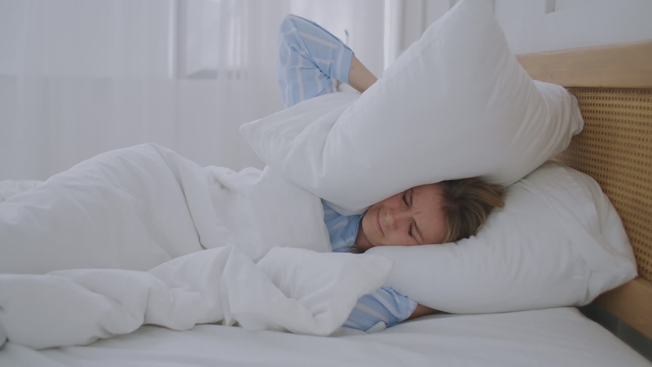 잠을 자려고 하는 젊은 짜증나는 여자 나쁜 시끄러운 방에 짜증나는 이웃들이 베개로 귀를  ⁇ 는 화난 소녀 학생 큰 소음으로 방해받는 문제 침대에 누워 깨어나 불면증으로 고통받습니다