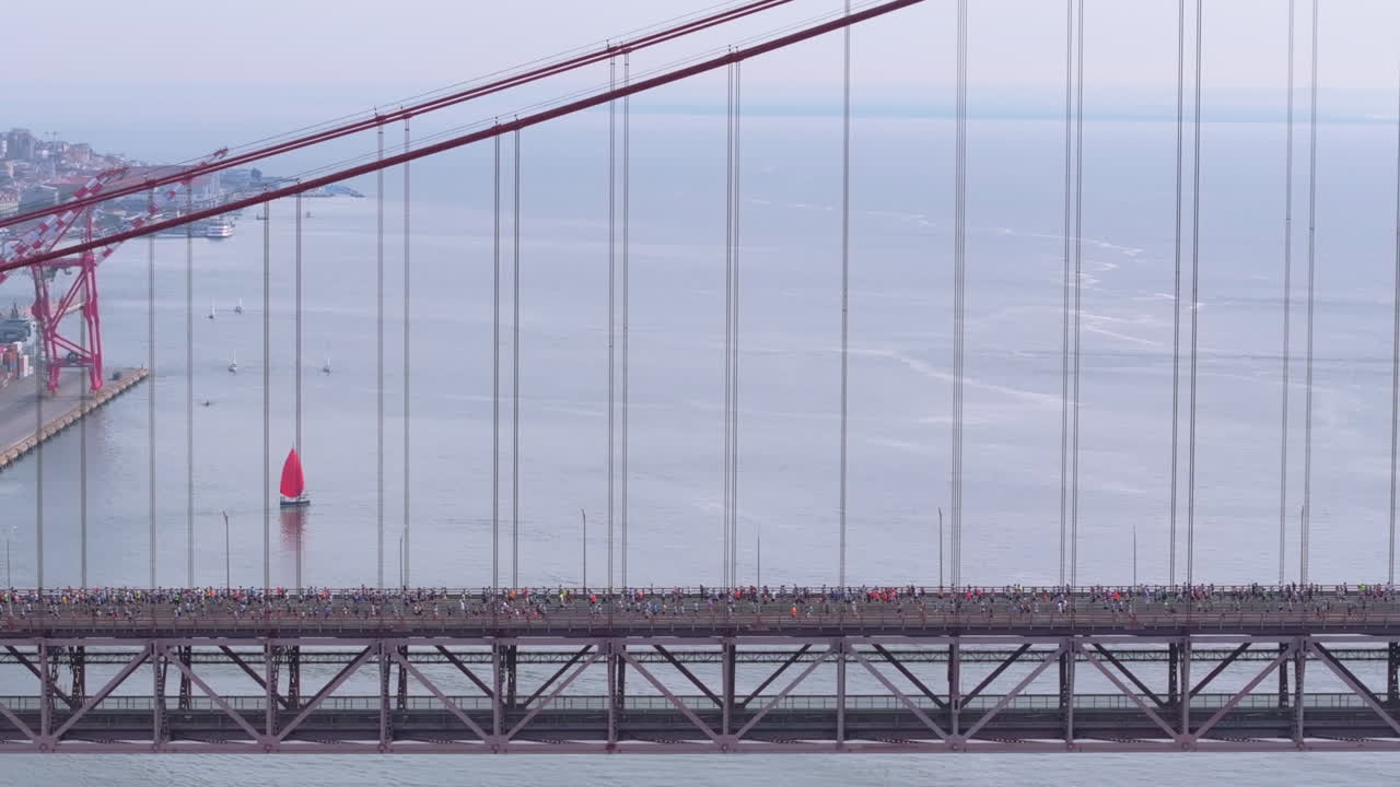 Side-on aerial drone shot of half marathon and 10K long-distance running event in Lisbon, Portugal, Europe. Runners crossing the iconic famous red 25th April bridge