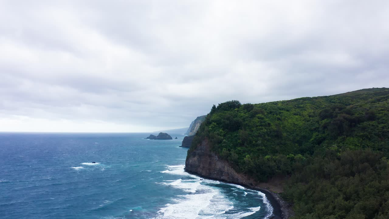 Steep green cliffs of Polulu Valley in Hawaii rise over turquoise ocean waves that roll gently toward the dark shoreline, while a blanket of thick clouds drifts overhead in this wild landscape.