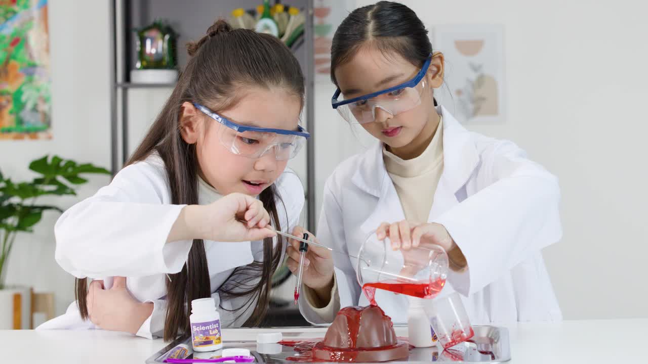 Dua gadis muda Asia berjas lab dan berkacamata pelindung menuangkan cairan merah ke dalam model gunung berapi.