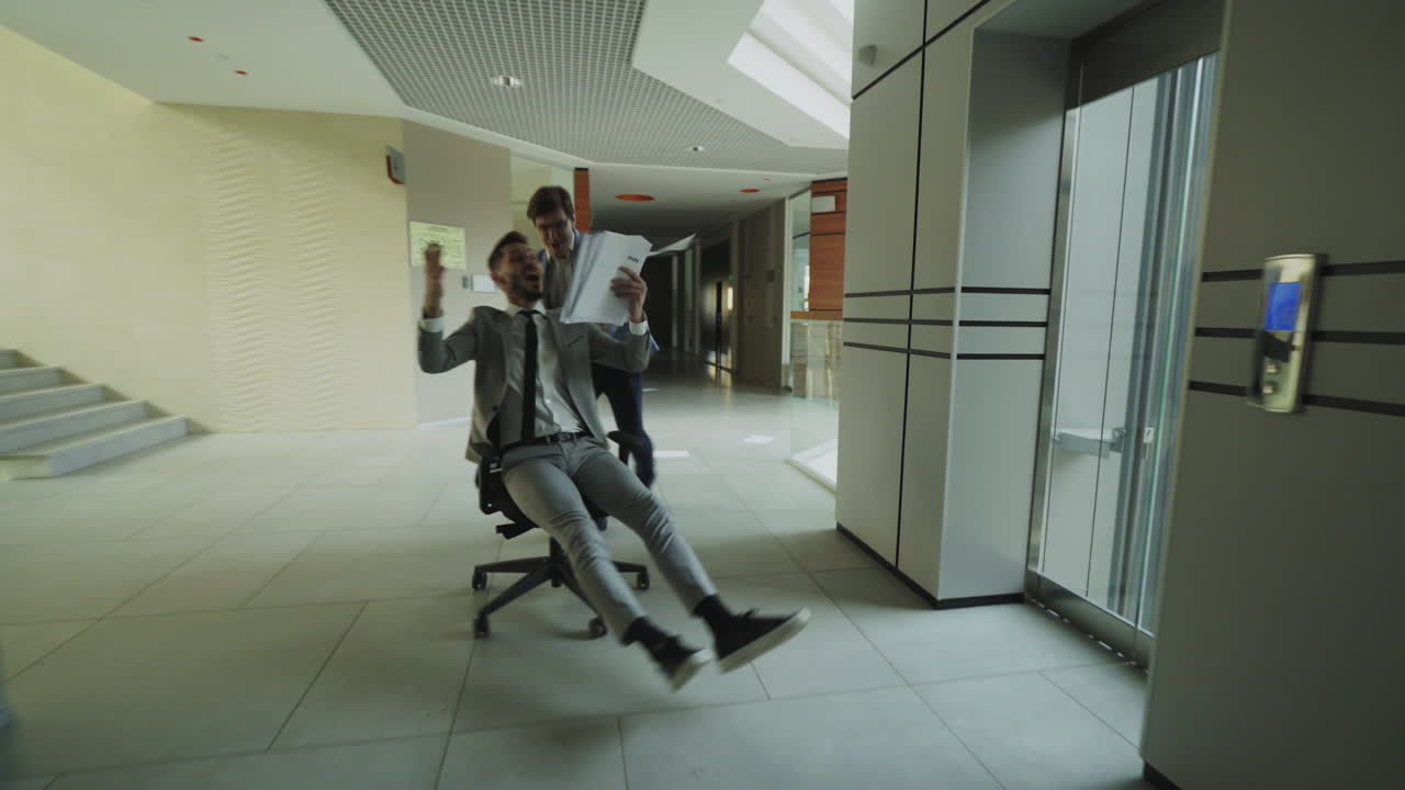 Businessmen Riding Chairs in Office Hallway