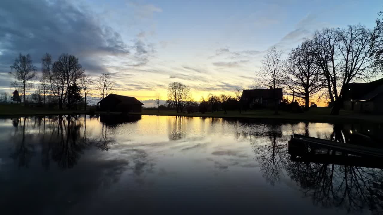 Silhouettes of farmlands as sun sets over calm pond with reflections of trees and clouds in peaceful countryside setting