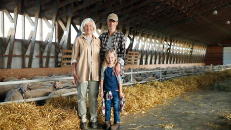 Portrait-of-Caucasian-grandmother,-mother-and-little-girl-smiling-at-camera-in-a-stable-with-sheep-flock