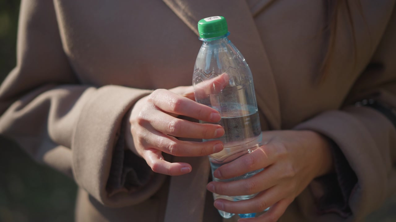 Close-up wanita memegang dan dengan lembut menggenggam botol air plastik bening bertutup hijau dalam cahaya alami yang lembut. Subjek mengenakan mantel krem, tangan terlihat dengan kuku yang rapi, dan latar belakang.