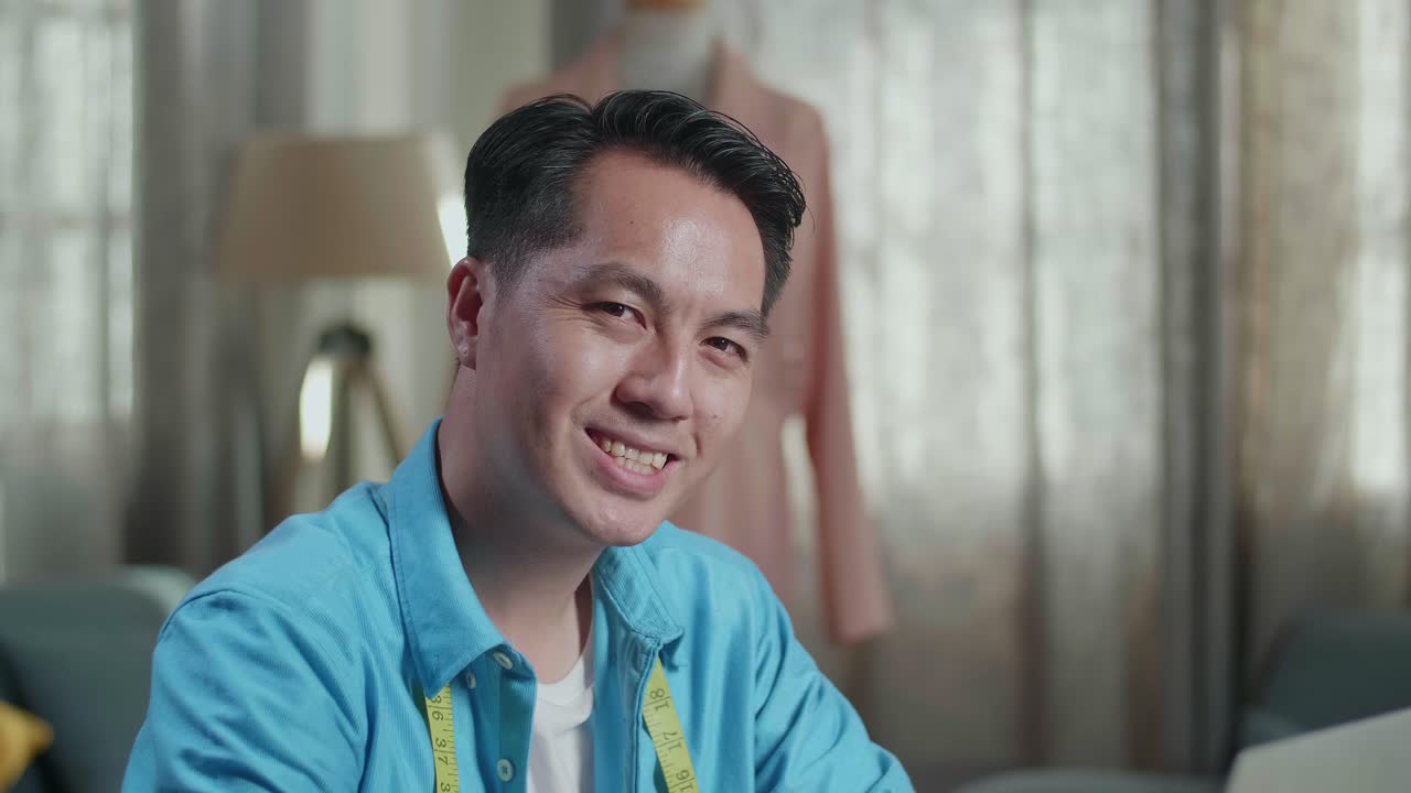 스튜디오에서 옷을 디자인하는 동안 노트북에서 일하고 카메라에 미소 짓는 아시아 남성 디자이너의 클로즈업