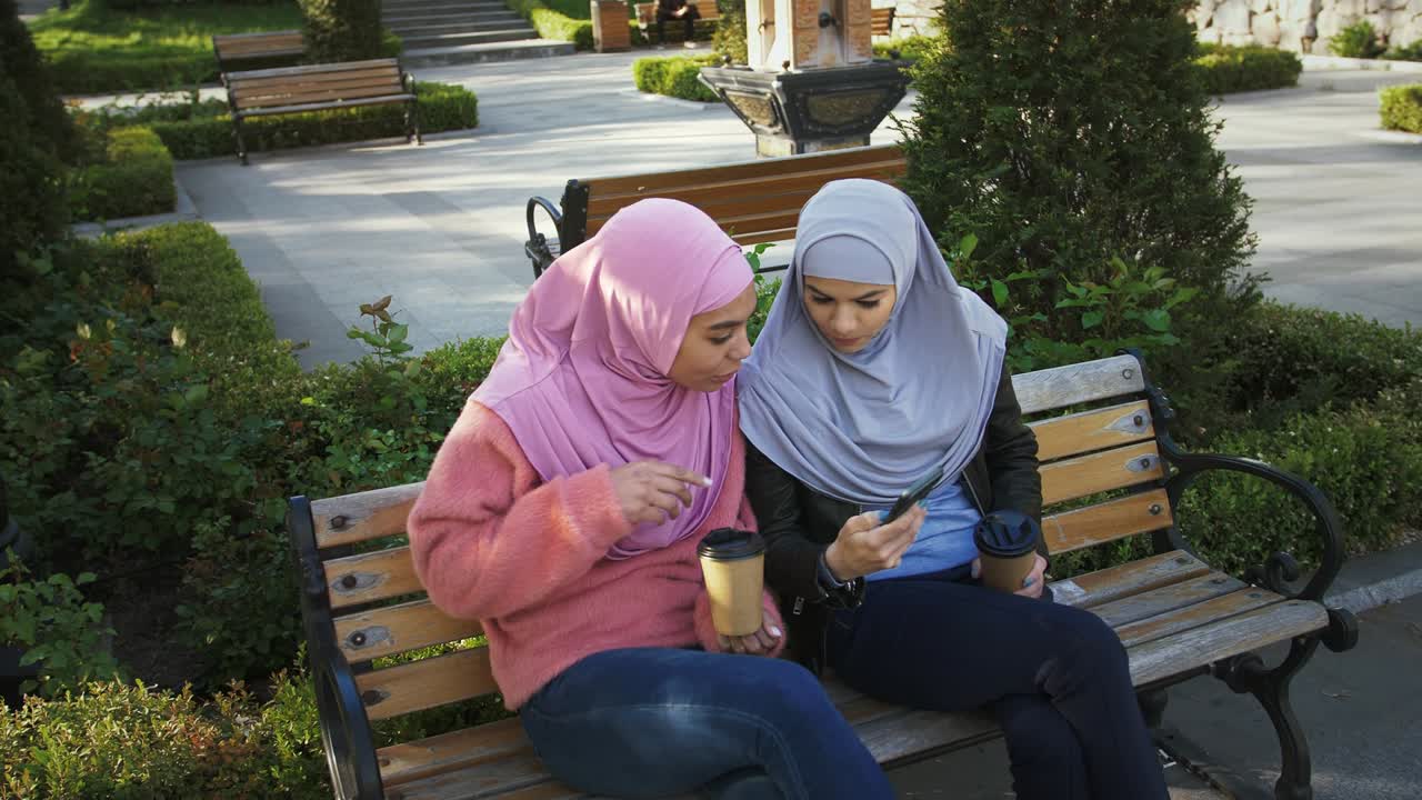 dos chicas musulmanas con trajes casuales, hijabs coloridos. disfrutando de café, mirando la pantalla de su teléfono inteligente. sentadas en un banco en el parque.