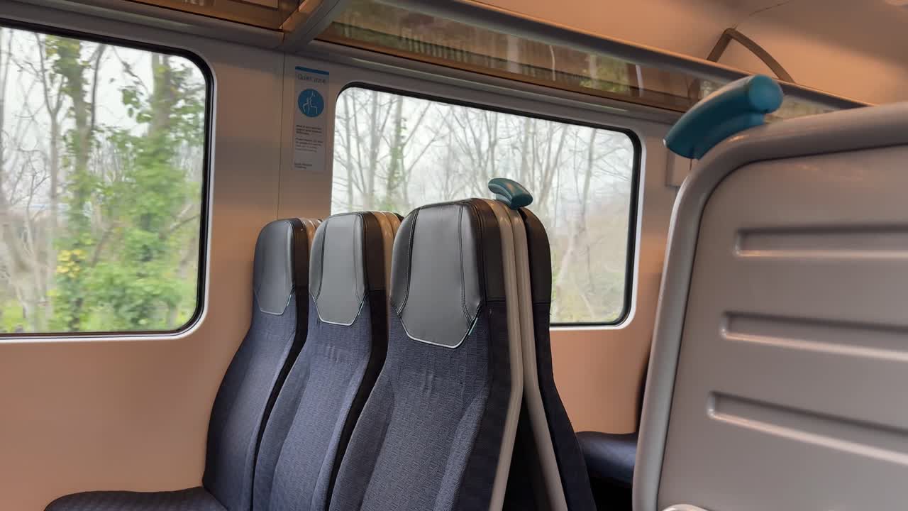 L'interno di un treno in movimento mostra sedili vuoti e una vista di alberi e paesaggio che scorrono fuori dal finestrino in una giornata nuvolosa