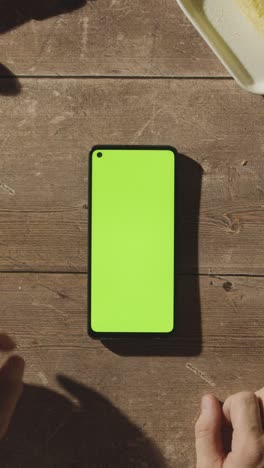 Toma-De-Video-Vertical-De-Una-Persona-Que-Usa-Un-Teléfono-Móvil-De-Pantalla-Verde-En-Una-Mesa-De-Madera