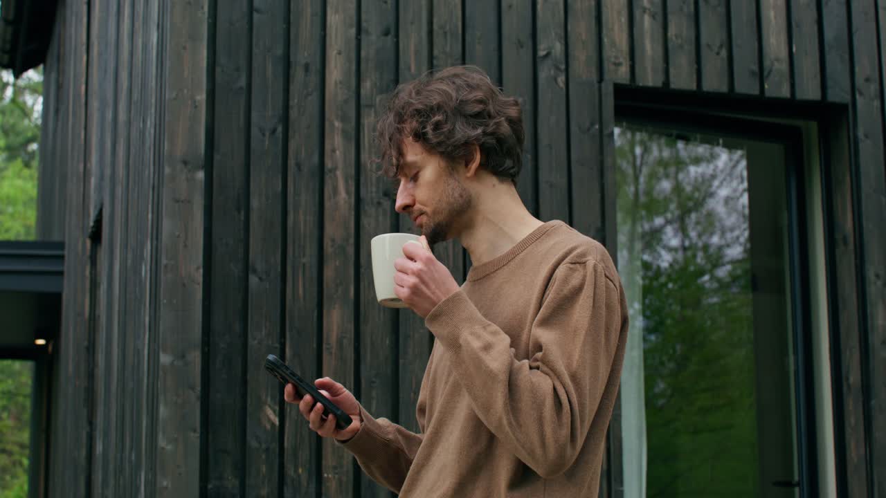 현대적인 오두막 밖에서 커피를 마시고 전화를 사용하는 남자