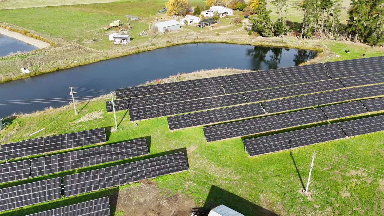Drone footage captures a solar farm amidst lush greenery and a reflective pond under bright daylight in Cromwell, New Zealand