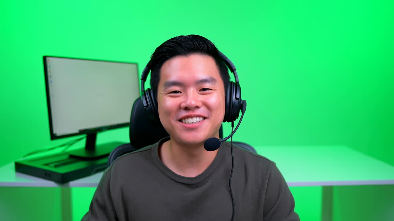 Uomo asiatico sorridente con cuffie in uno studio verde, seduto alla scrivania accanto al monitor, mentre parla al microfono e indossa abiti casual.