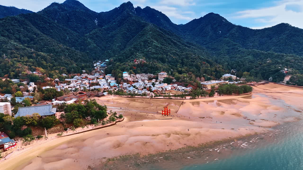 Sandy shore of the Inland Sea at the Miyajima island, Japan. People walk near the iconic Grand Torii Gate at Itsukushima Shrine. View on the multiple houses dwelling at the foot of green mountains.