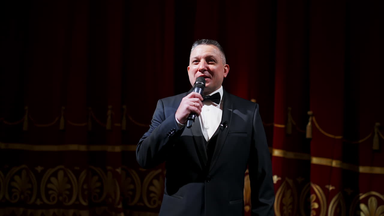 Male announcer with microphone on the stage. Actor in black suit on red scene curtains background.