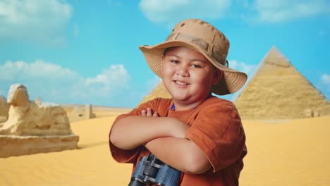 asian boy with a hat and binoculars crossing arms and smiling to camera while traveling in giza pyramid. boy researcher examines something, travel adventure, close up side view