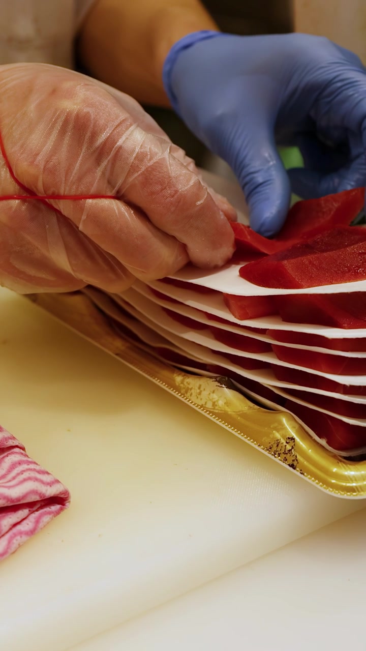 Close-up shot of a chef in blue gloves carefully layering raw tuna onto paper
