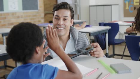 Happy-diverse-male-teacher-with-tablet-helping-schoolchildren-in-classroom-at-elementary-school