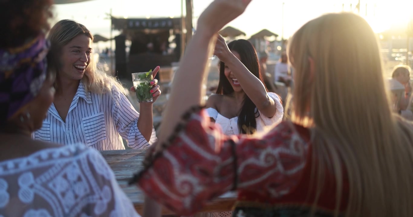 Glade, forskellige veninder nyder drinks på strandbar ved solnedgang