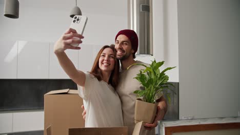 Happy-brunette-girl-in-a-white-t-shirt-hugging-her-brunette-boyfriend-in-a-red-hat-and-cream-t-shirt-who-is-holding-a-house-plant-and-taking-a-selfie-with-her-girlfriend-in-her-new-modern-apartment-after-moving-with-boxes-around