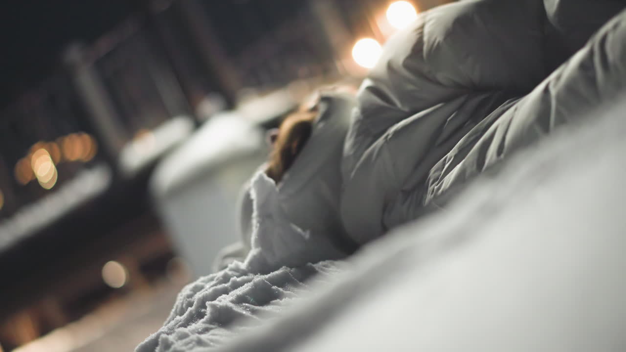 Side view of young girl in winter jacket lying on snow-covered ground swinging arms gently while surrounded by soft bokeh lights and snowy atmosphere in park at night, creating serene