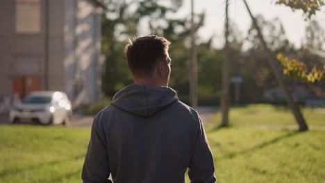 back view of young man in gray hoodie walking through outdoor grassy area in front of blurred building on sunny day; serene, introspective moment with relaxed, casual attire captured from behind